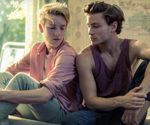 Film Still DIE MITTE DER WELT, Romanverfilmung von Jakob M. Erwa, mit Louis Hofmann, Svenja Jung und Sabine Timoteo; Phil und Nicholas sitzen zurückhaltend, aber sich zugeneigt auf dem Bett