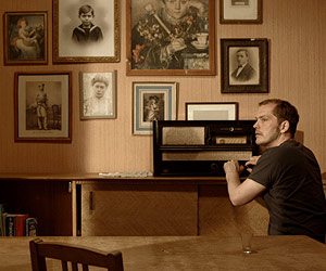 Film Still Liebmann von Jules Herrmann; Antek Liebmann (gespielt von Godehard Giese) sitzt vor einem Radio und einer Wand mit vielen alten Fotos