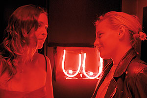 Film Still BELOW HER MOUTH von April Mullen; Jasmine (gespielt von Natalie Krill) und Dallas (gespielt von Erika Linder) begegnen sich in einer Bar vor einer roten Lichtinstallation weiblicher Brüste
