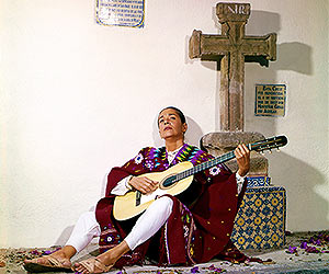 Film Still CHAVELA von Catherine Gund und Daresha Kyi über die lateinamerikanische Ranchera-Sängerin Chavela Vargas mit Pedro Almodóvar; Chavela sitzt im Poncho vor einem Steinkreuz und spielt Gitarre