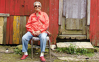 Film Still CHAVELA von Catherine Gund und Daresha Kyi über die lateinamerikanische Ranchera-Sängerin Chavela Vargas mit Pedro Almodóvar; Chavela sitzt mit Sonnenbrille vor einer Holzhütte