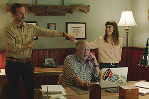 Film Still EXTRA TERRESTRES – EXTRA TERRESTRIALS von Regisseurin Carla Cavina mit Elba Escobar; Teresa (gespielt von Marisé Alvarez) gibt ihrem Bruder Junior (gespielt von Yamil Collazo) einen Fist-Bump hinter dem Rücken ihres Vaters Arcadio (gespielt von Sunshine Logroño), der an seinem Schreibtisch sitzt