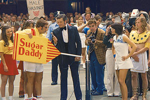 Film Still BATTLE OF THE SEXES – GEGEN JEDE REGEL von den Regisseuren Jonathan Dayton & Valerie Faris mit Bill Pullman, Alan Cumming, Elisabeth Shue und Sarah Silverman; Billy Riggs (gespielt von Steve Carell) steht in einer Tennis-Arena neben Wimbledon-Ikone Billie Jean King (gespielt von Emma Stone) und hält ein Schild mit der Aufschrift Sugar Daddy