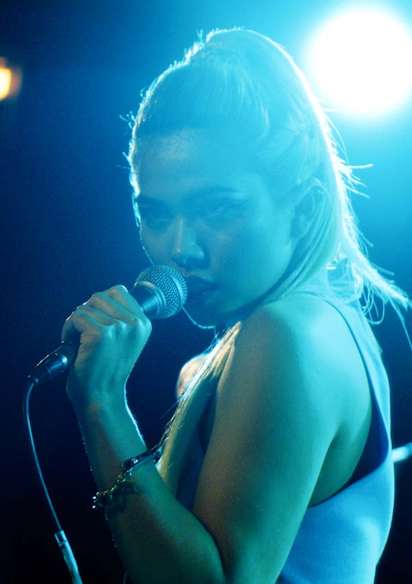 Film Still BECKS von den Regisseuren und Ko-Autoren Daniel Powell und Elizabeth Rohrbaugh mit Lena Hall, Christine Lahti, Dan Fogler und Mena Suvari; Sängerin Lucy (Hayley Kiyoko) singt in hellem, blauen Scheinwerferlicht ins Mikrofon