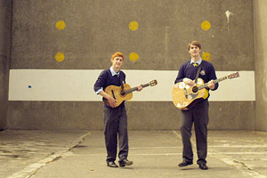 Film Still HANDSOME DEVIL von John Butler; der rothaarige Ned (gespielt von Fionn O'Shea) und sein Zimmergenosse Conor Masters (gespielt von Nicholas Galitzine) stehen mit Gitarren auf einem Sportplatz aus brutalem Sichtbeton