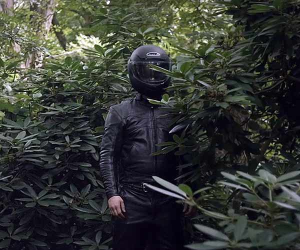 Film Still JAHR DES TIGERS – THE YEAR I LOST MY MIND, ein queerer Berlin-Thriller von Tor Iben mit Alexander Tsypilev, Julien Lickert, Patrick G. Boll, Michael del Coco und Christian Harting; ein mysteriöser Mann steht in schwarzer Motorradmontur aus Leder sowie geschlossenem Visier seines schwarzen Helms zwischen grünen Büschen