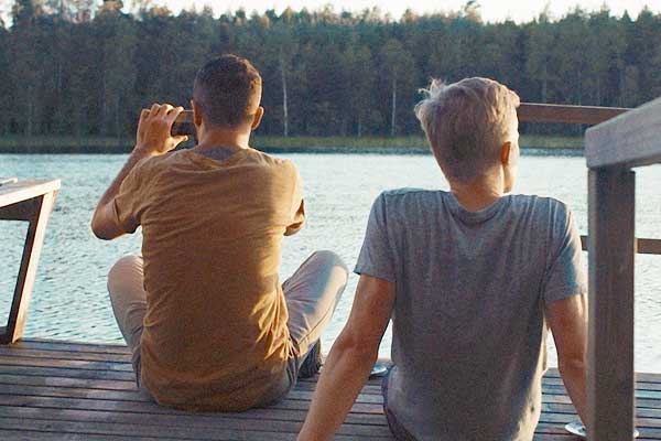 Film Still DIE HÜTTE AM SEE – A MOMENT IN THE REEDS, eine schwule Romanze zwischen einem Finnen und einem syrischen Flüchtling von Filmemacher Mikko Makela mit Janne Puustinen, Boodi Kabbani und Mika Melender; Leevi (gespielt von Janne Puustinen) sitzt mit Tareq (Boodi Kabbani), der mit seinem Smartphone ein Foto der Aussicht macht, auf einem Holzsteg am See