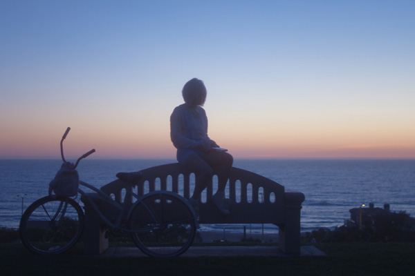 Film Still DADDY ISSUES von Regisseurin und Cutterin Amara Cash aus den USA, 2018; Maya Mitchell (gespielt von Madison Lawlor) sitzt neben ihrem Fahrrad auf einer Bank vor einem Ozean-Ausblick und einem spektakulären Sonnenuntergang