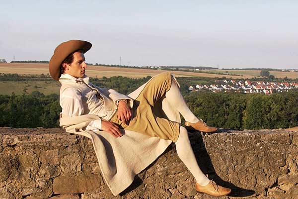 Film Still MÄNNERFREUNDSCHAFTEN von Regisseur Rosa von Praunheim über die Freundschaften unter Weimarer Schriftstellern; ein Schauspieler sitzt in historischem Kostüm und Hut auf einer Mauer auf einer Anhöhe vor einem Dorf im Wald