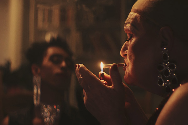 Film Still TUCKED von Autor und Regisseur Jamie Patterson aus England, 2018; die Drag-Queen Jackie (gespielt von Derren Nesbitt) zündet sich ohne Perücke eine Zigarette an und Faith (gespielt von Jordan Stephens) steht mit Diadem und großen Ohrringen im Hintergrund